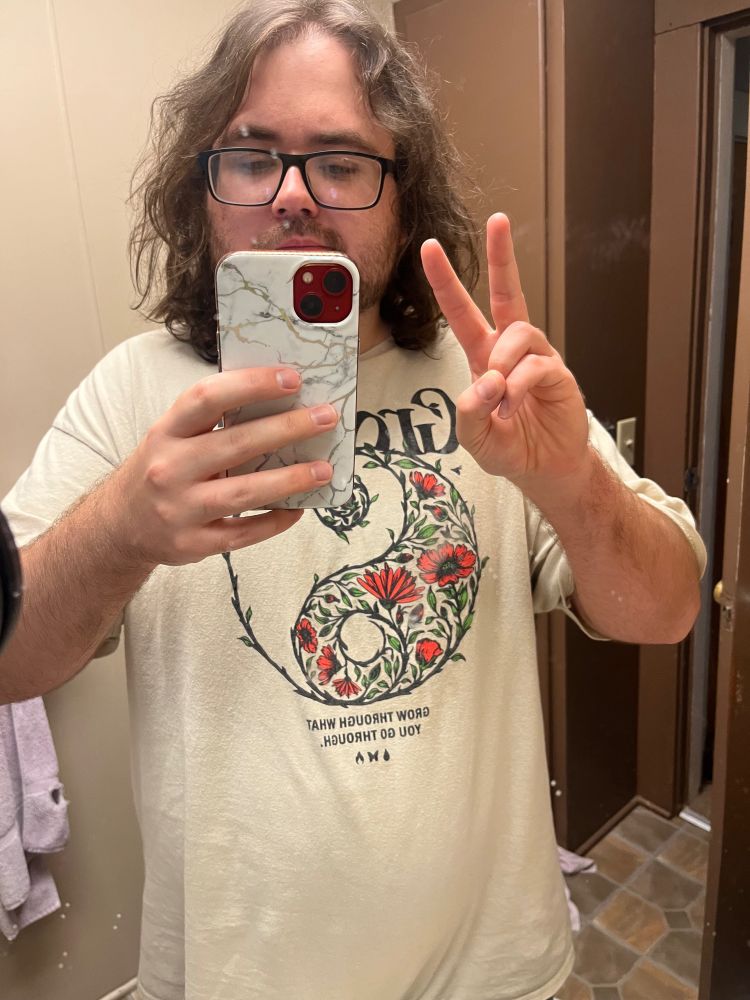 Me giving a peace sign while holding my phone in the mirror. 

I’m wearing a beige shirt with a ying/yang symbol made of flowery plants that says “Growth, Grow through what you go through” 