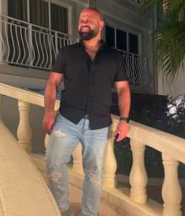 Bald man with a black beard, torn jeans, black shirt, and a watch on his right arm