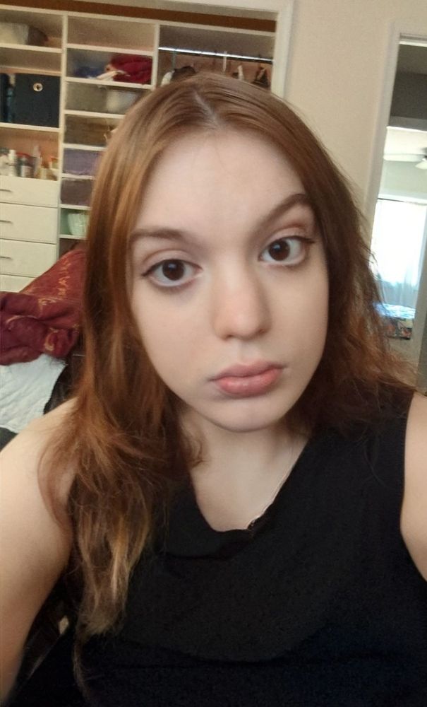 A woman with long, brown hair and a neutral expression takes a selfie in a room with shelves filled with assorted items in the background while wearing black dress