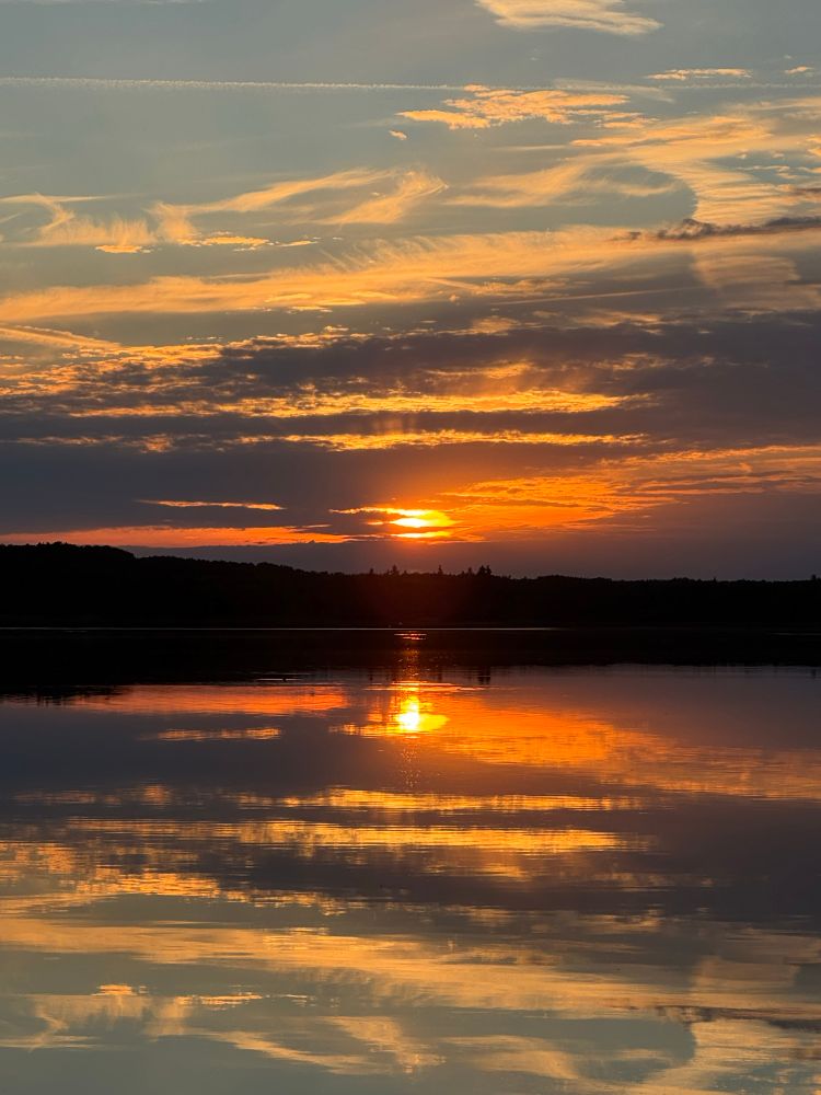 A sun setting behind clouds casting an orange glow in the sky and reflections in the still waters in front of it. 