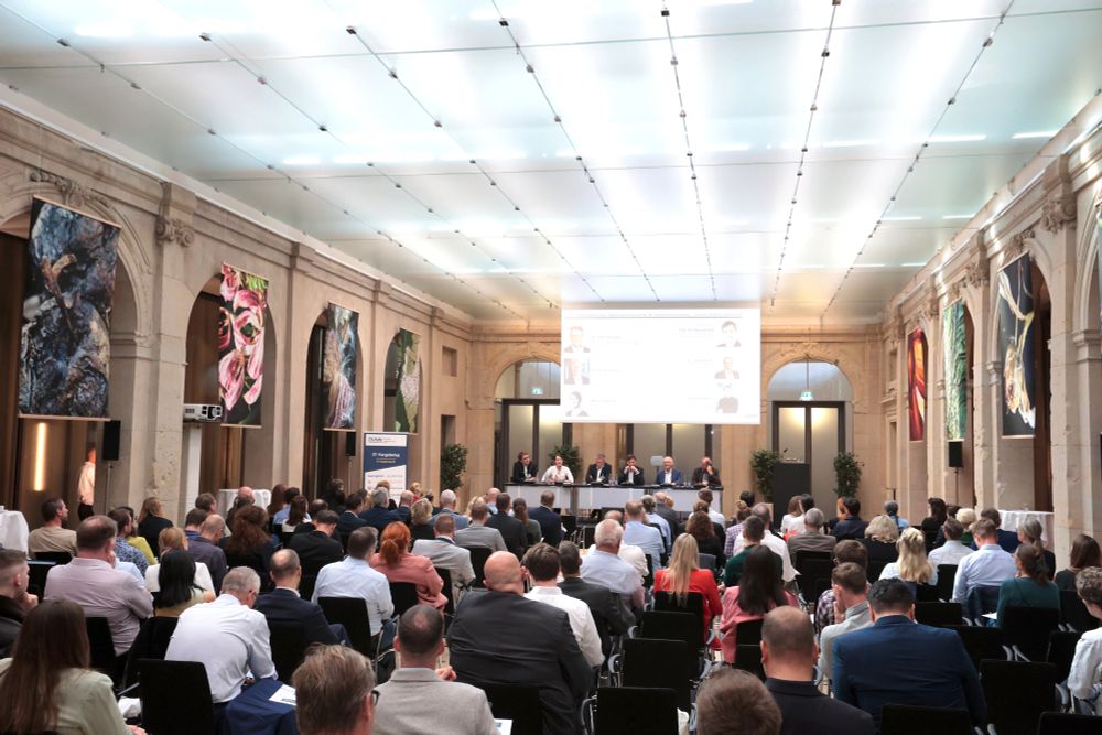 Weitwinkel-Aufnahme vom hinteren Ende eines Konferenzraums. Vorne sitzen die sechs Panelteilnehmenden an einem Tisch, davor sieht man das Publikum von hinten, das auf das Panel schaut.