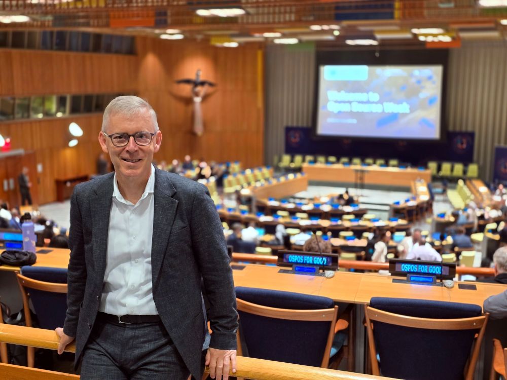 Peter Ganten lehnt an einem Geländer und schaut freundlich in die Kamera, hinter ihm sieht man einen großen Plenarsaal im Gebäude der Vereinten Nationen, auf einem Bildschirm an der Rückwand des Saales steht in verschwommener Schrift „Welcome to Open Source Week“