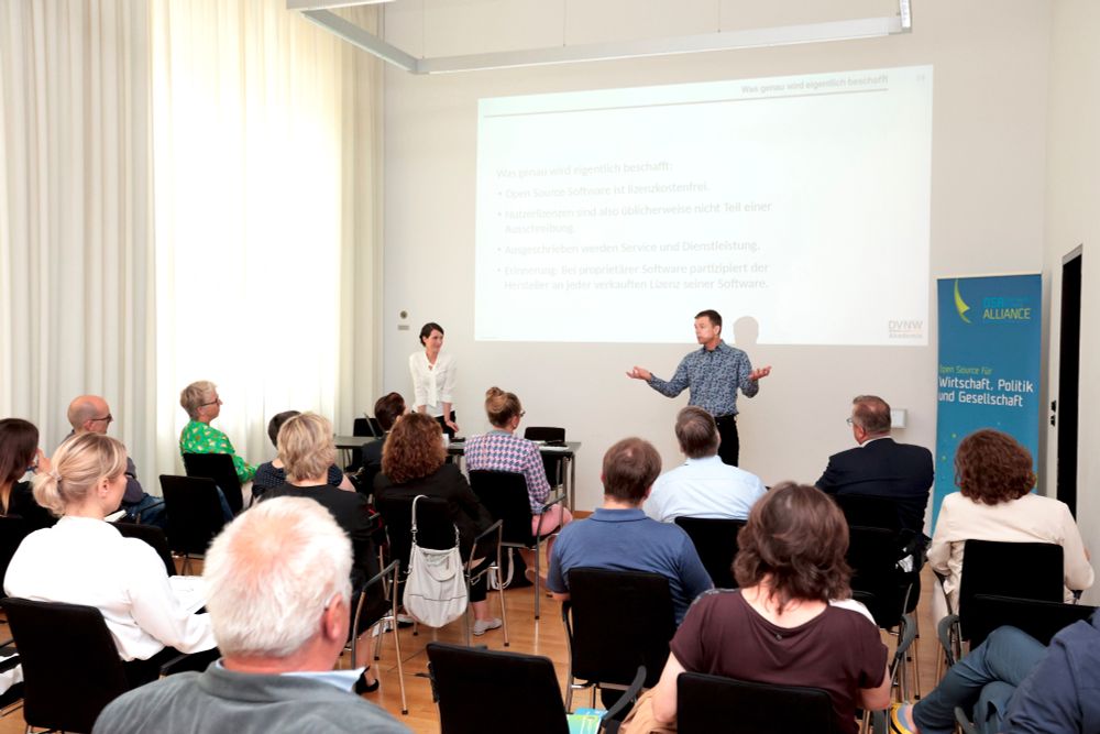 Von hinten aus einem Workshopraum über die Köpfe der Teilnehmenden fotografiert: Vorne stehen Miriam Seyffarth und Claus Wickinghoff, rechts neben ihnen ein Rollup der Open Source Business Alliance, auf dem &quot;Open Source für Wirtschaft, Politik und Gesellschaft&quot; steht. An der Wand im Hintergrund die Präsentation der beiden Speaker:innen.
