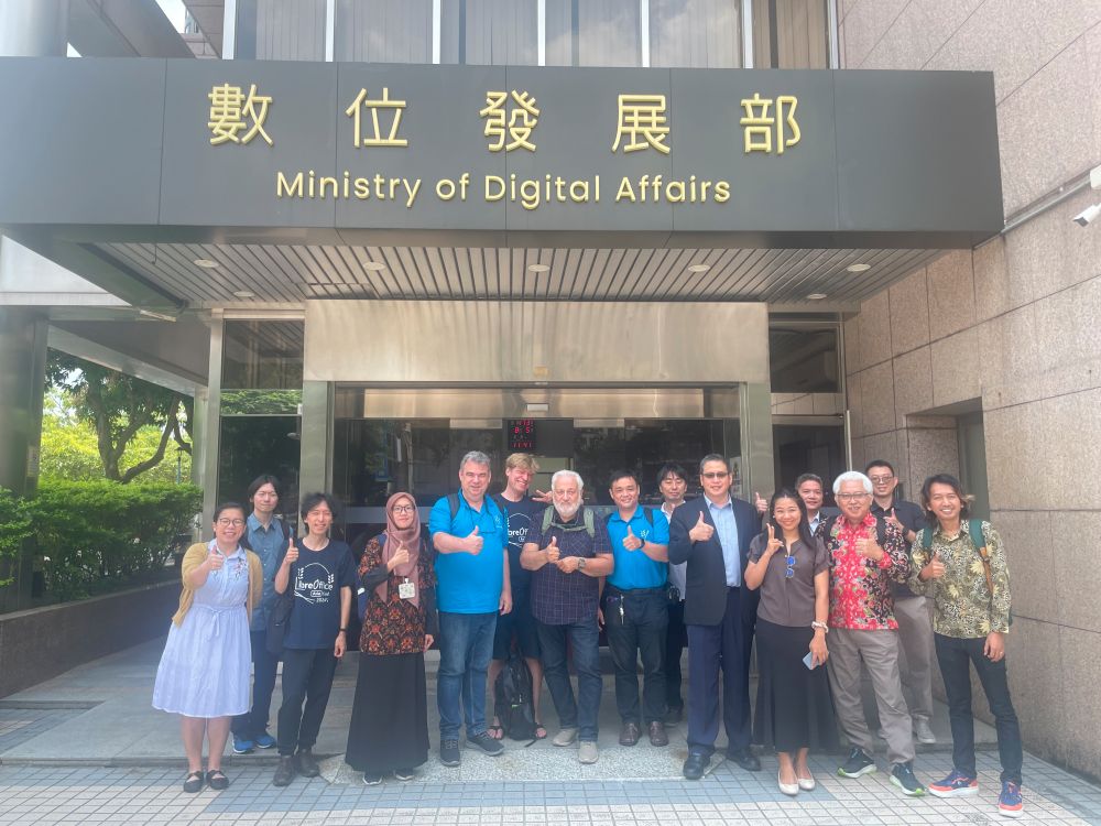 Eine Gruppe von 15 Personen steht vor dem Eingang des „Ministry of Digital Affairs“ in Taiwan. Über dem Eingang sind große Schriftzeichen in traditionellem Chinesisch sowie die englische Bezeichnung der Behörde angebracht. Die Personen – darunter Mitglieder der LibreOffice-Community und Delegierte der The Document Foundation – lächeln in die Kamera und zeigen Daumen-hoch-Gesten. Einige tragen LibreOffice-T-Shirts. Es ist ein sonniger Tag, im Hintergrund sind Glasfassaden und Grünpflanzen zu sehen.