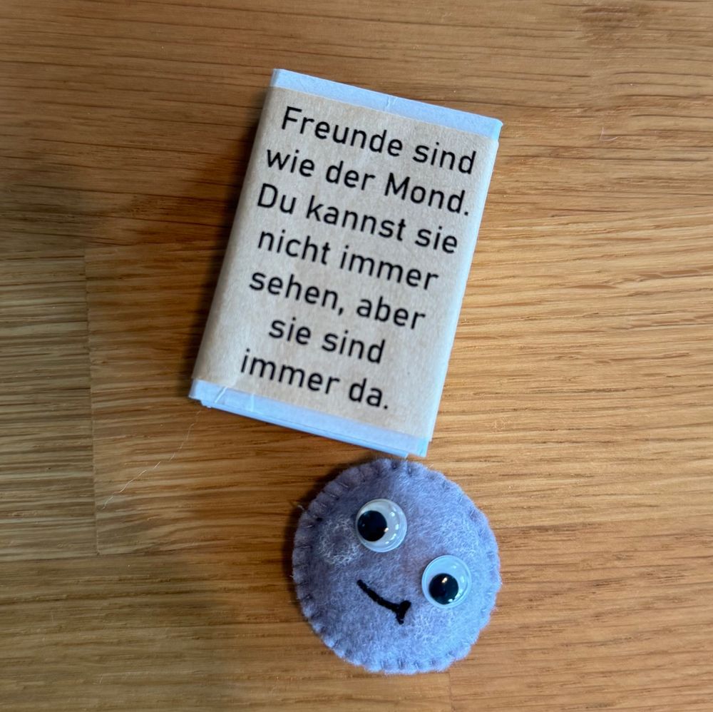 An inspirational quote about friendship is printed on a small book-like object. Next to it, there is a round, gray felt figure with googly eyes and a smiling face. The background is a wooden surface.