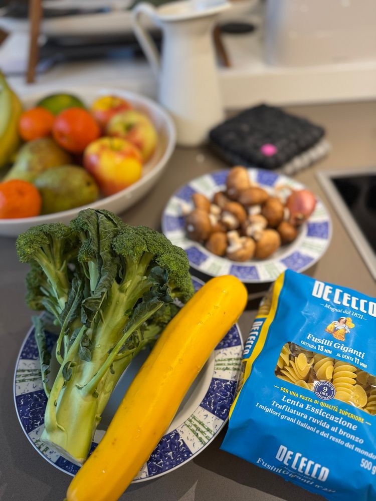 A collection of fresh vegetables, including broccoli and yellow zucchini, is placed on a decorative plate. In the background, there is a bowl of assorted fruits and a plate with mushrooms. A bag of De Cecco fusilli pasta is also visible, indicating