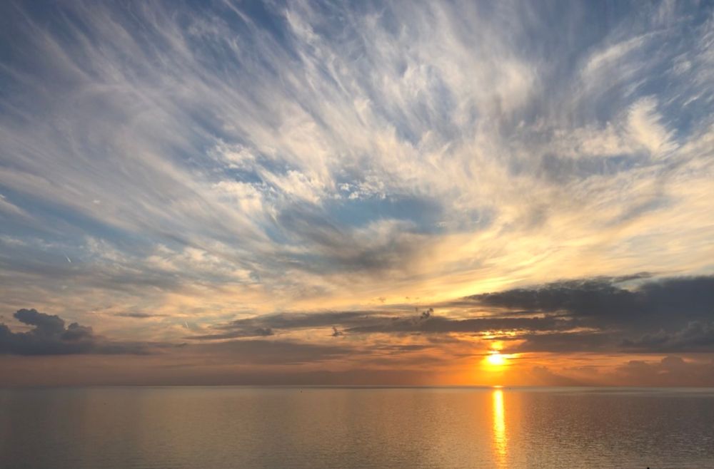 A serene sunset over a calm sea, featuring vibrant orange and yellow hues reflecting on the water, with wispy clouds scattered across a blue sky.