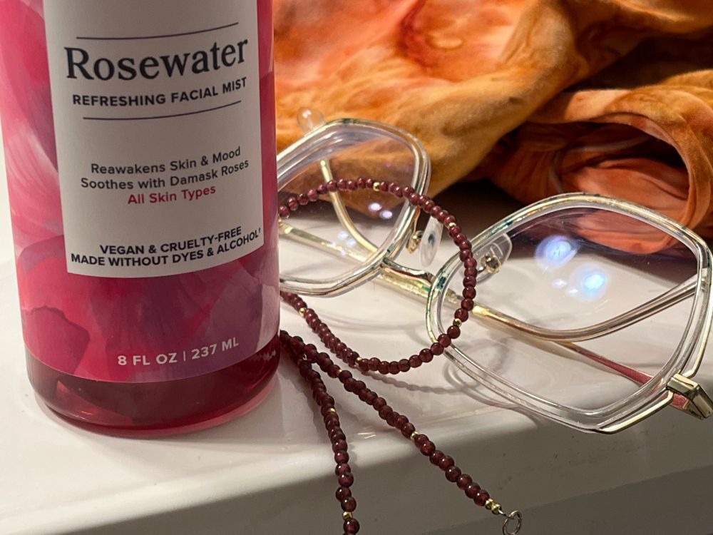 A bottle of rose water toner sits on a brightly lit countertop with a choker necklace made of small garnet beads, a pair of gold rimmed glasses, and a mottled orange and brown bandana. 