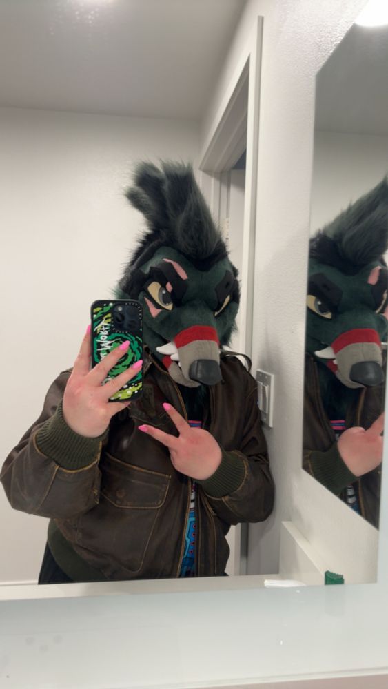 A mirror selfie of person wearing a vintage air force jacket and a snarling dark green wolf fursuit head. And doing a peace sign, showing off bubblegum pink fingernails 