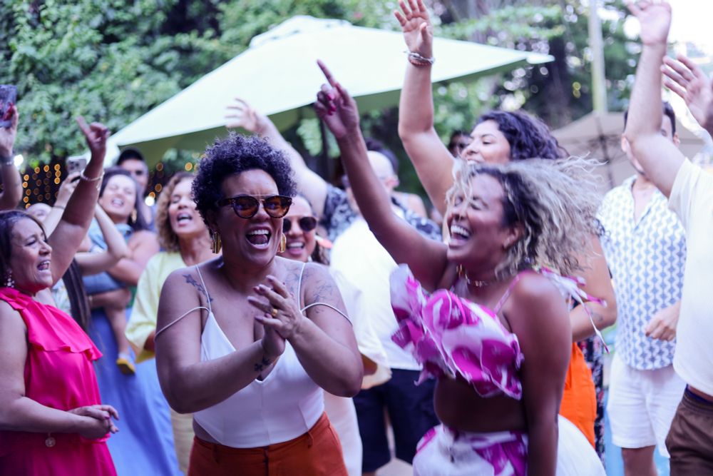Duas mulheres negras se divertindo e dançando. Uma delas sorri e bate palmas. A outra pula com o bracinho pra cima e sorri, o movimento é tão rápido que ela sai ligeiramente desfocada 