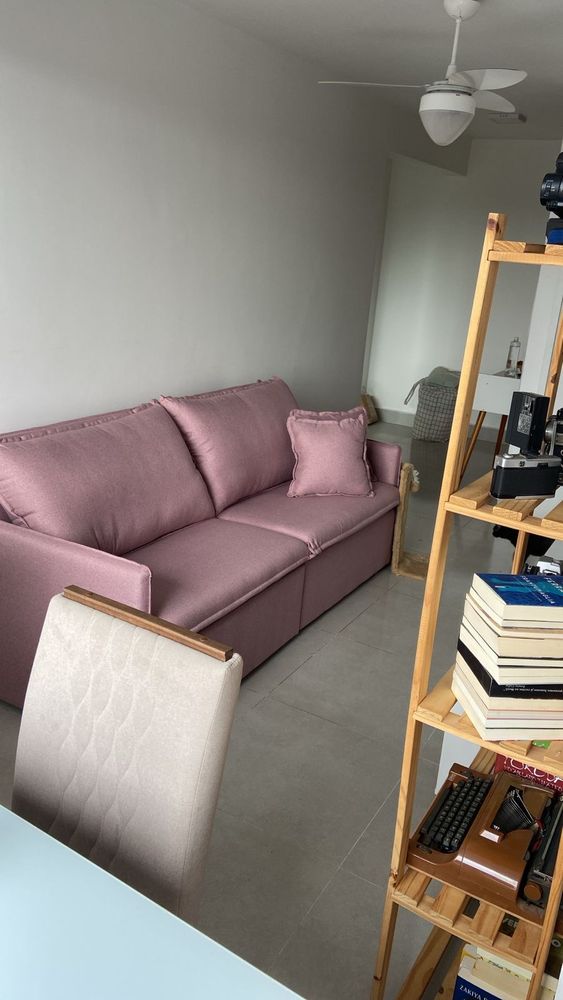 Meu sofá rosa, 3 lugares, reclinável, uma delícia de confortável. Do lado direito da foto tem uma estante de madeira com livros, uma máquina de escrever.