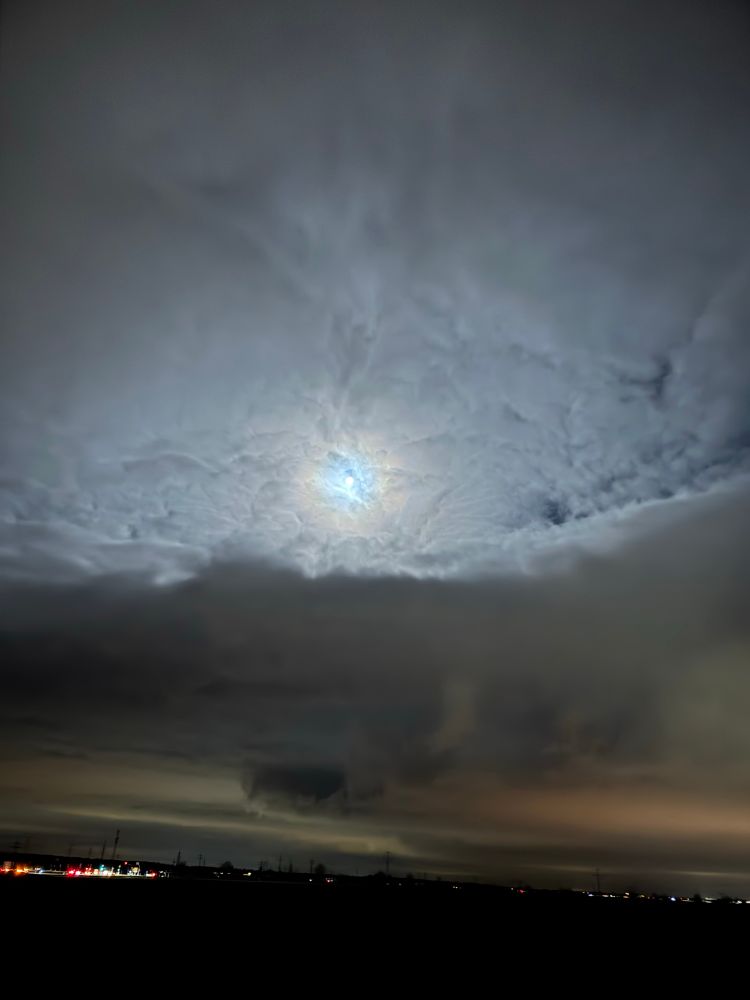 Das Bild zeigt eine dramatische Winterhimmelslandschaft kurz vor Sonnenaufgang, aufgenommen vor 7:00 Uhr morgens. Der Vollmond ist hinter dichten, teils zerrissenen Wolken zu sehen und strahlt schwaches Licht aus, das die Wolkendecke erhellt. Am unteren Bildrand ist eine dunkle, wolkenverhangene Szenerie zu erkennen, während im Hintergrund die Lichter einer Stadt oder eines Dorfes sichtbar sind. Die Farbtöne des Himmels variieren von Dunkelgrau bis hin zu leichtem Blau, das von der Mondbeleuchtung hervorgehoben wird.
