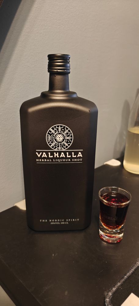 Photo of a bottle of Valhalla Herbal Liquor Shot, with a shot glass on the side filled with clear brown liquor from the bottle