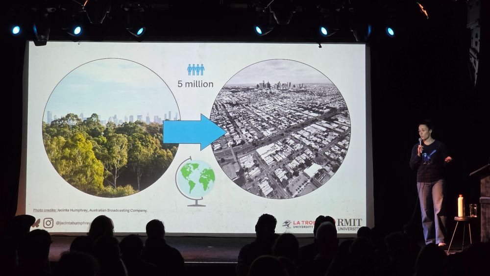 Dr Jacinta Humphrey stands off the right-hand-side of the stage, beside a large projection of her slides. On the screen is a circular photo of a leafy, green city with a blue arrow pointing towards a greyscale photo of roads, houses, shops and concrete as far as the eye can see. These images indicate the current transition we are seeing in Melbourne.