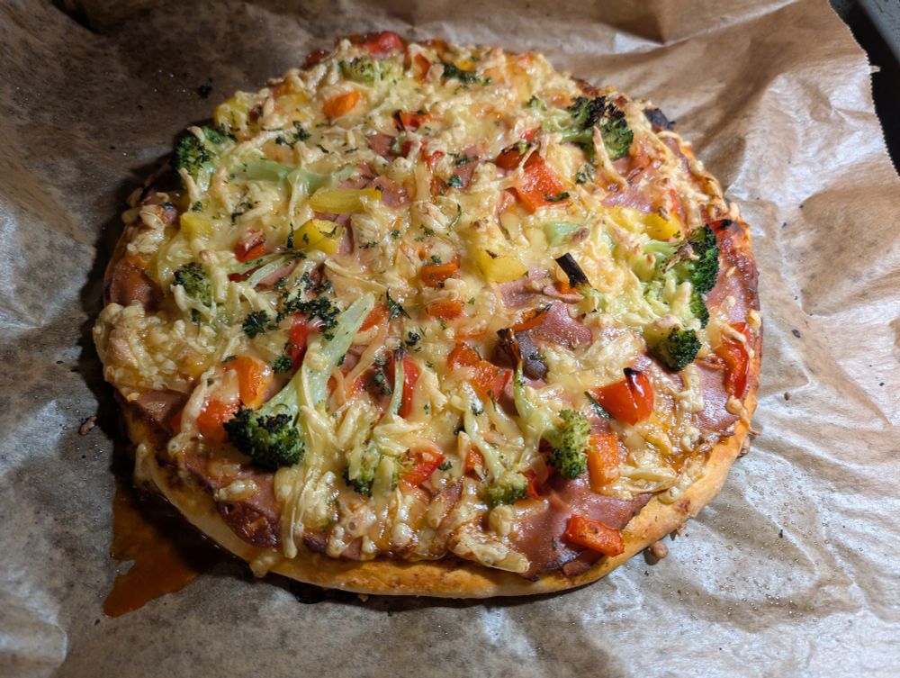 Eine plantbased Pizza mit Schinken, Brokkoli, Paprika, Knoblauch und ordentlich Keese liegt auf Backpapier. Dier Ansicht ist von oben. 
Das Backpapier ist ezwas verknuddeld.