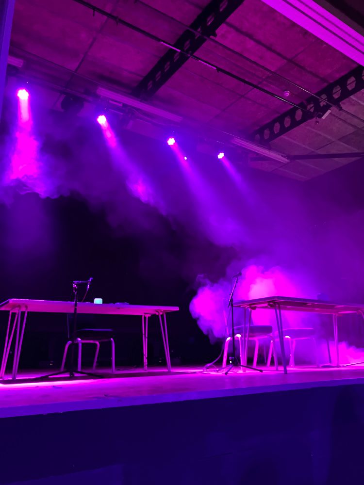 Photo of a stage with two tables, one with one chair and one with two chairs. Lit with purple and pink light and probably a bit too much fog machine/atmosphere
