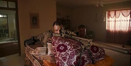 American solider Elliot Miller (played by Cosmo Jarvis) in a seized Iraqi apartment aims sniper rifle out of a window. Rifle is propped up by mattress and blankets. Miller is flanked by a second soldier.