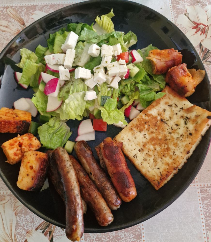 Salat mit Kräuter Schafskäse, Grillkäse, Berner Würstchen, Nürnberger Würstchen und marinierten Grillkäsewürfeln. Auf blauem Teller. Hübsche Wachstischdecke. 