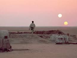 The binary sunset Scene from Star Wars: A New Hope. The Lars homestead bunker is in the foreground, and the sands stretch far into the distance. In the foreground centre, Luke skywalker, back to the camera is watching the sunset. The white sun is high, the orange one low, as they head toward the horizon.