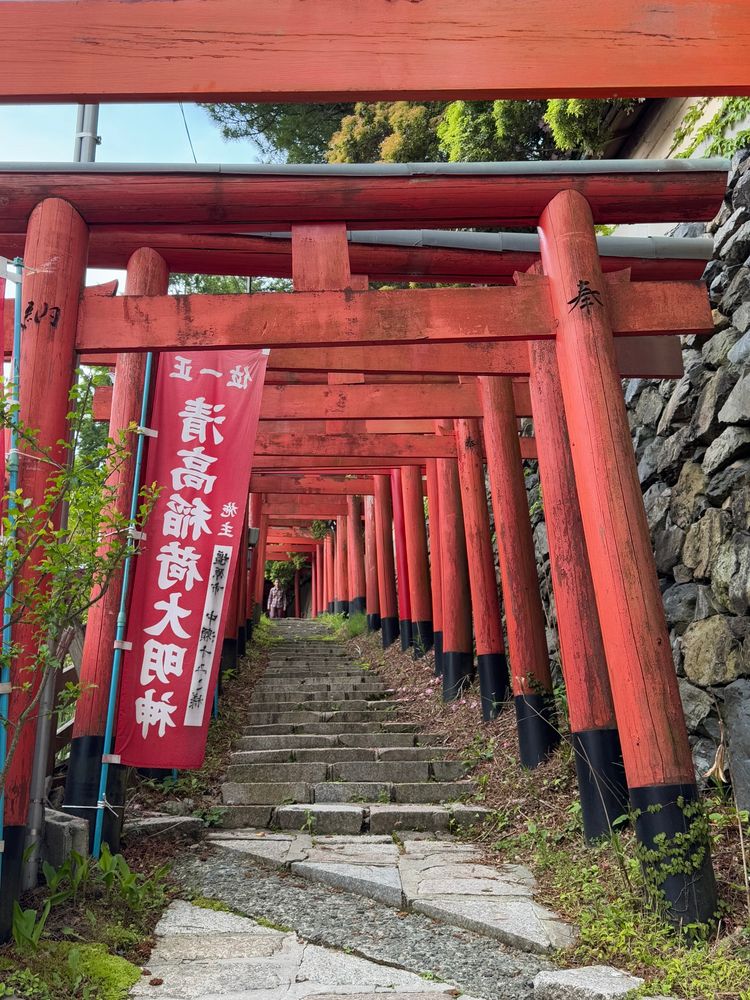 Rote Tore ( Torii) stehen hintereinander. Darunter führt leicht ansteigend eine Natursteintreppe nach oben. Rechts im Bild sieht man Teil einer Natursteinmauer und links im Bild eine rote Fahne mit japanischen Buchstaben.