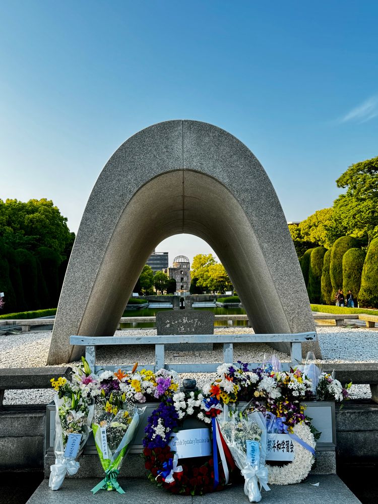 Blick durch grauen Betonbogen, in dem ein Gedenkstein steht, im Hintergrund brennt die ewige Fackel und dahinter steht die Ruine des Atombombendoms. Vorne im Bild stehen Blumenssträusse und Trauerkränze.