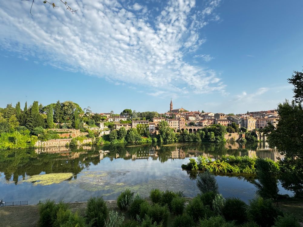 Im Bild schauen wir von der anderen Seite eines Flusses auf ein am Ufer liegendes mediterranes Stadtteil . Welches sich auf etwa mittlerer Bildhöhe erstreckt. Dieses spiegelt sich, zusammen mit dem blauen Himmel im Klaren Flussbett wieder.