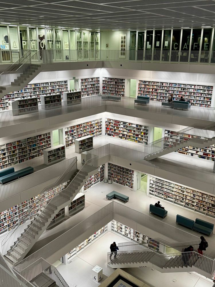 Blick von dem oberen Stockwerk in die Galerie einer Stadtbücherei. Alles in weiß gehalten, auch die Treppen, zu den darunter liegenden Stockwerken, vor dem Bücherregalen stehen türkisfarbene Sitzgelegenheiten.