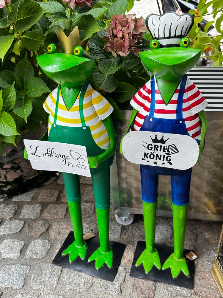 Zwei stehende grüne Metall - Frösche mit Ringelshirt und Latzhose. Linker Frosch trägt gelb geringeltes Shirt mit grüner Latzhose und Schild, auf dem „Lieblings Platz“steht, auf dem Kopf trägt er eine goldene Krone. Der re. Frosch trägt ein rot Weiss geringeltes Shirt und eine blaue Latzhose und hält ein Schild mit „Grillkönig“. Auf dem Kopf sieht man eine weiße Kochmütze.