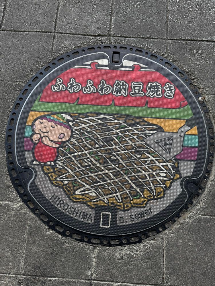 Gullydeckel, auf dem ein japanischer Pfannkuchen mit Bratenwender abgebildet sind. Links auf dem Pfannkuchen steht noch ein kleines Männchen mit freudigem Gesicht. Unten auf dem Deckel steht Hiroshima.