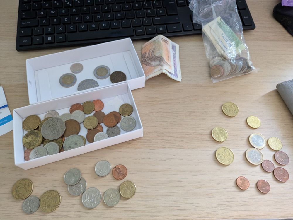 Various coins and notes spread out on a desk, some in a small shallow box. Various currencies, not a clue which is which.