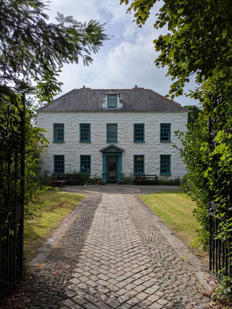 Through black metal gates and beyond trees and bushes, a white two storey building in the distance, the trims of the windows and front door are turquoise. Front door in the centre, two portrait rectangle windows evenly spaced either side of it, five windows all evenly spaced on the second storey and one window centrally placed embedded in the slate roof. A grey brick path leads to the building. The morning sky above the building is pale blue dotted  with a few clouds.