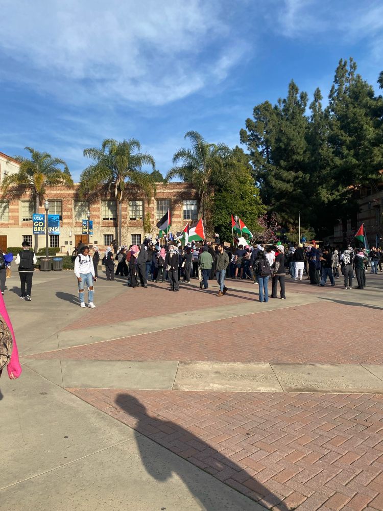 UCLA Bruin quad. Students for Justice in Palestine in with Palestinian flags. Lots of red brick on the ground with a brick building and palm trees in the background.