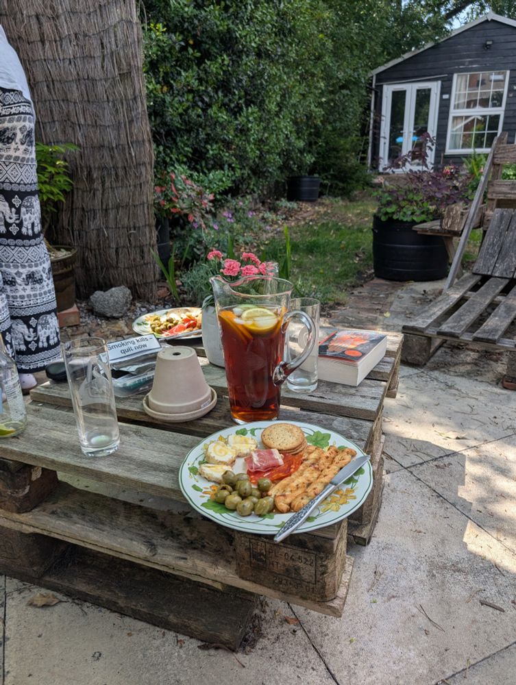Outdoor pallet wood table with plates of antipasto, a jug of Pimm's, and a book, on it