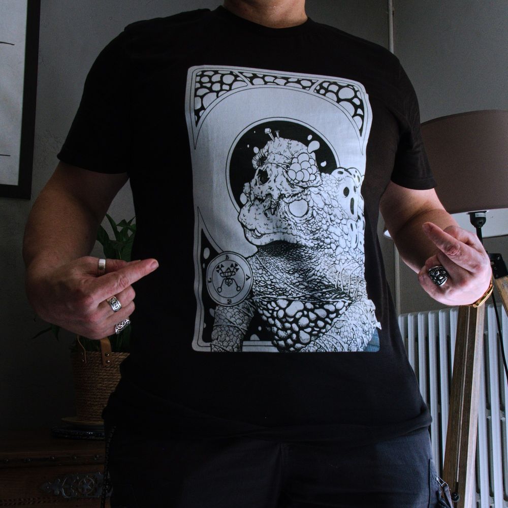 Photo d'une personne portant un t-shirt floqué d'un dessin du démon Baël représenté par un crapaud aux yeux blancs, le corps et les bras enroulés de fils, portant un crâne de chat sur la tête, percé de deux clous et dont les orbites suintent d'un liquide étrange. Son dos est recouvert de têtes hurlantes. En arrière plan un cadre formant un cercle central, en haut des motifs arrondis, en bas des bulles de liquide sur fond noir. Son sceau est visible en bas à droite