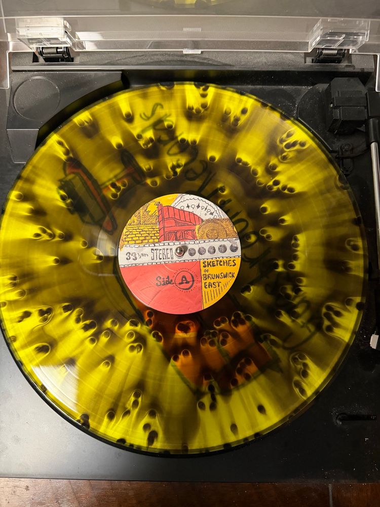 The vinyl of King Gizzard & The Lizard Wizard with Mild High Club’s album Sketches of Brunswick East sitting on a turntable. The vinyl itself is a yellow and black ice cloudy vinyl, otherwise referred to as migraine edition wax, which means a yellow and black splatter on the record. Almost looks like peacock feathers.