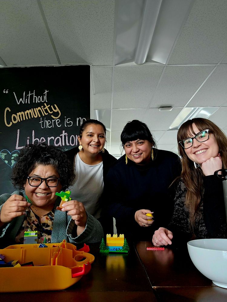 Photo includes four people in a collaborative office space. One individual is holding LEGOs, while all four smile. Two people are standing (center) and two are sitting. An Audre Lourde quote is in the background, which reads, "Without community there is no liberation." A white bowl sits on the right, a yellow case is on the left. 