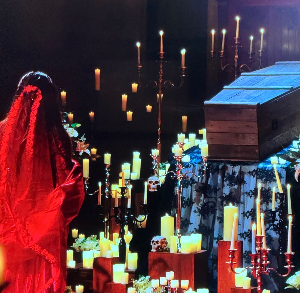 mega dramatic scene of a woman in red dress and veil facing a closed coffin and a hundred lit candles 