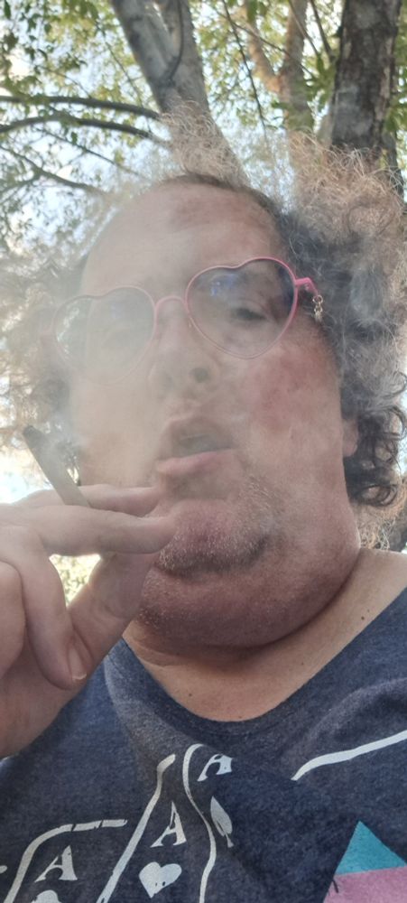 A large white trans femme woman in a cloud of smoke while holding the joint she's smoking. Her expression is one of dismissive uninterest.