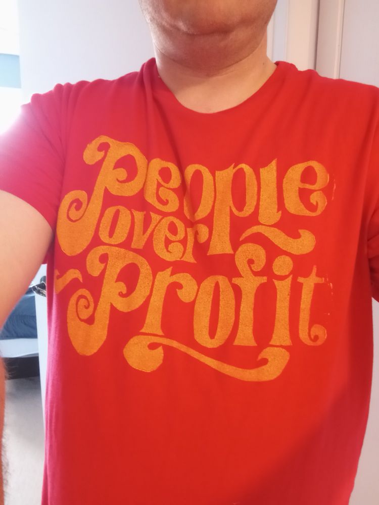 A man taking a selfie in a red shirt that says, "People Over Profit" in yellow lettering.