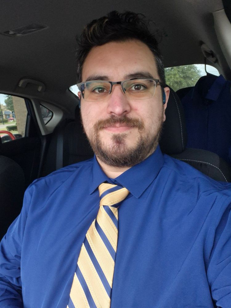 Taxtm in a blue button-up shirt, wearing a tie with gold and blue diagonal stripes.