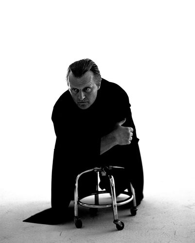 Black and white photo of Rutger Hauer leaning against a short rolling stool.