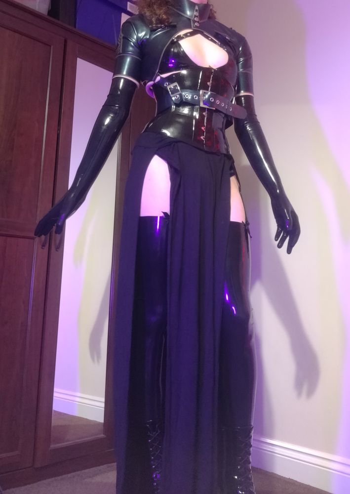 Standing tall in high heeled boots and latex stockings showing through a double split skirt. The Dolls waist is tightly cinched in by a corset as it wears a military style bolero and long gloves.