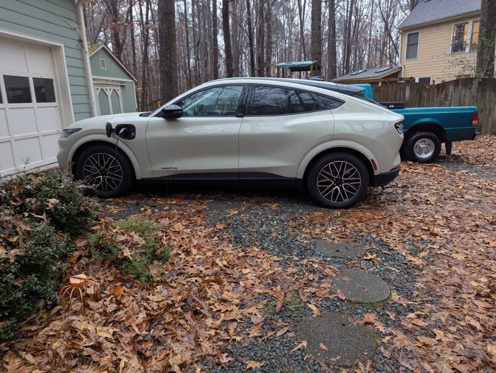 A light creamy beige Ford MachE plugged in next to a garage in a gravel driveway full of wet leaves