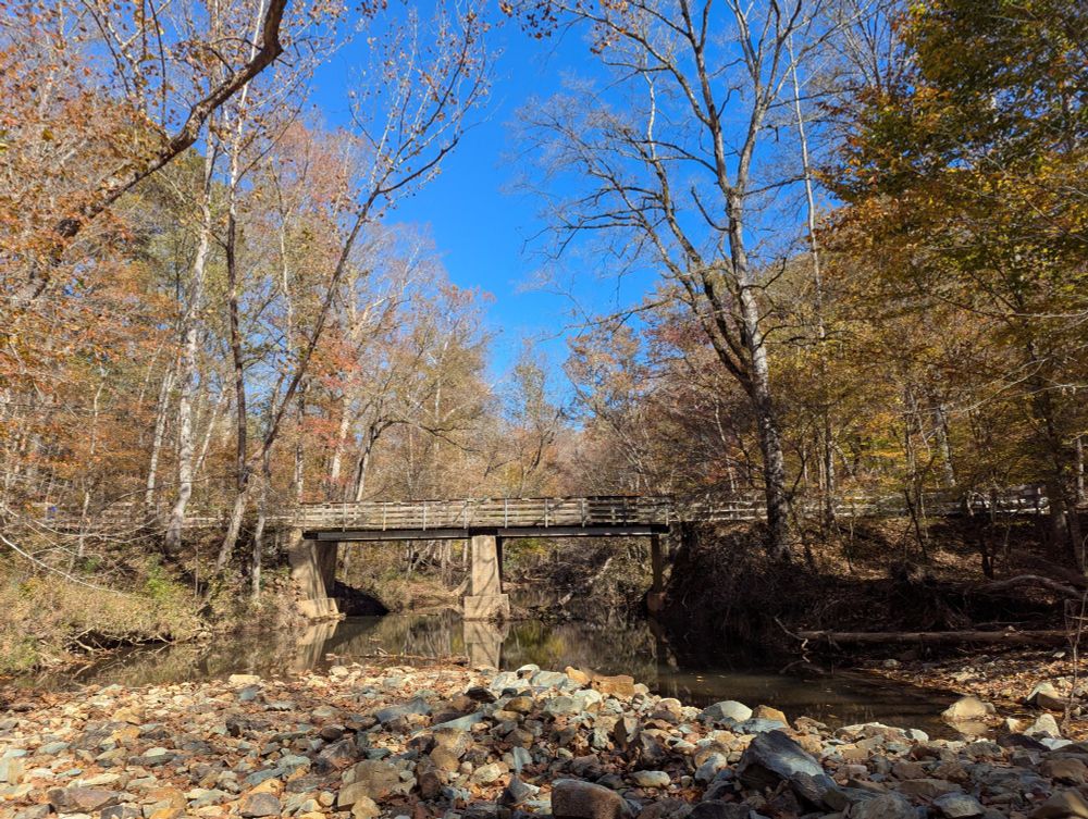 a wooden and concrete bridge crossing a rocky stream thru bright yellow and orange woods, some bare trees, mid day, blue sky