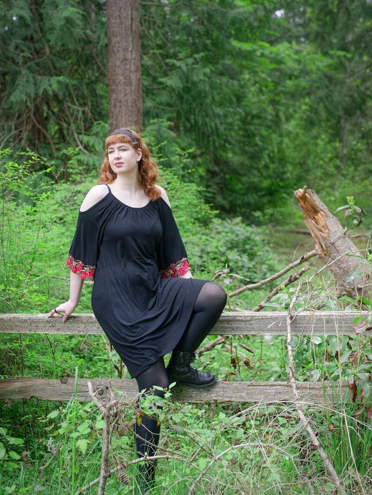 A red-haired person wearing a loose black dress with rose-embroidered sleeves and combat boots sits on a small fence in a lush Pacific Northwest forest.