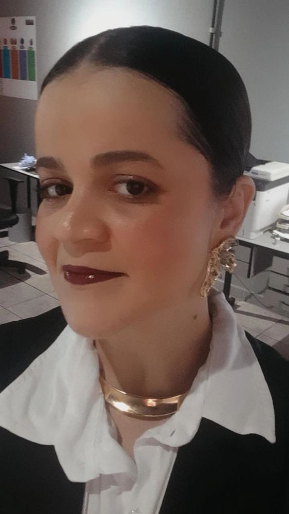 Selfie de uma mulher com o cabelo partido no meio em um coque pra trás. Blusa branca, colete preto e acessórios dourados.