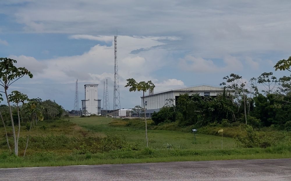 Das Ariane 6-Launchpad von einer Straße aus gesehen: Ein hohes Gebäude mit Torflügeln und vier Blitzableiter-Masten zu allen Seiten. Davor, näher zum Betrachter hin, die flache Halle, in der die Komponenten der Rakete zusammengefügt werden.