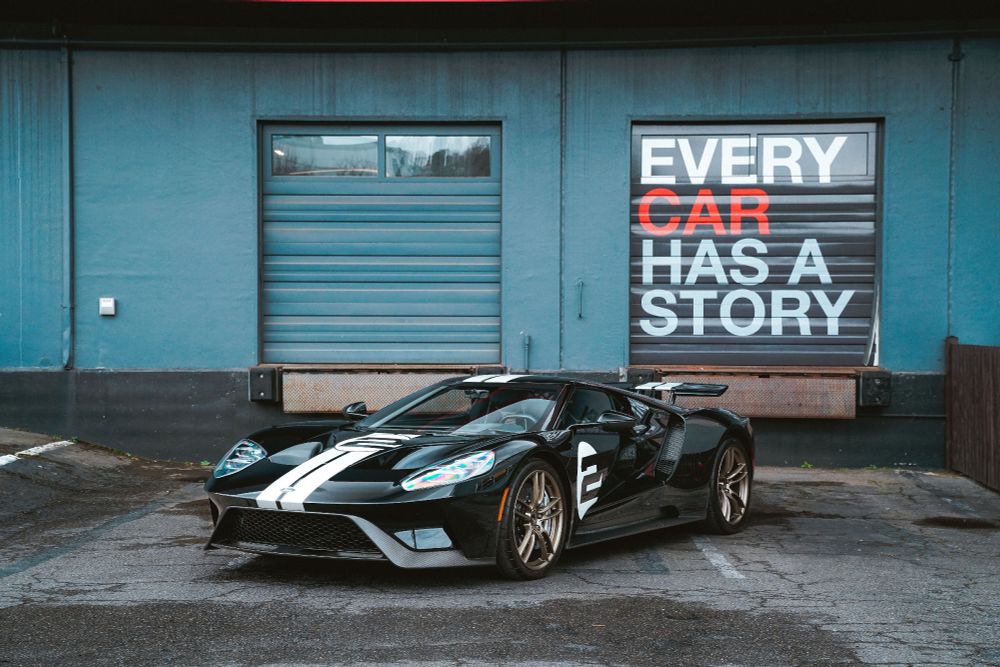 Image shows a very sleek black car, a 2017 Ford GT, with white decals and the number 2 painted on its side. The car is very shiny against the matt blue background of garage doors and a black road. Text on one of the garage doors says every car has a story.