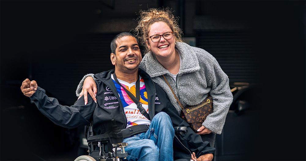 Two people smiling and embracing while posing for a photograph, one using a wheelchair, at a sunny outdoor event. 