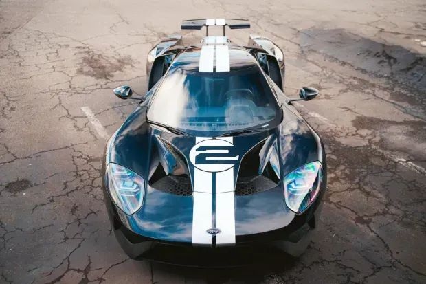 A front-on shot of a Ford GT on a cracked road. The natural lighting makes the headlight iridescent and shows the clouds overhead.